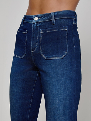 A close-up of a person wearing the Noya Cropped Slim Flare Jean, featuring a high-waisted fit, stretch dark blue denim with white stitching and front pockets. Only the lower torso and upper legs are shown.