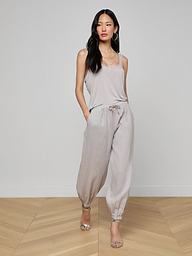 A woman stands against a plain white background, hands in pockets, wearing the Mariko Linen Jogger with a light beige sleeveless top, hoop earrings, and open-toe heels.