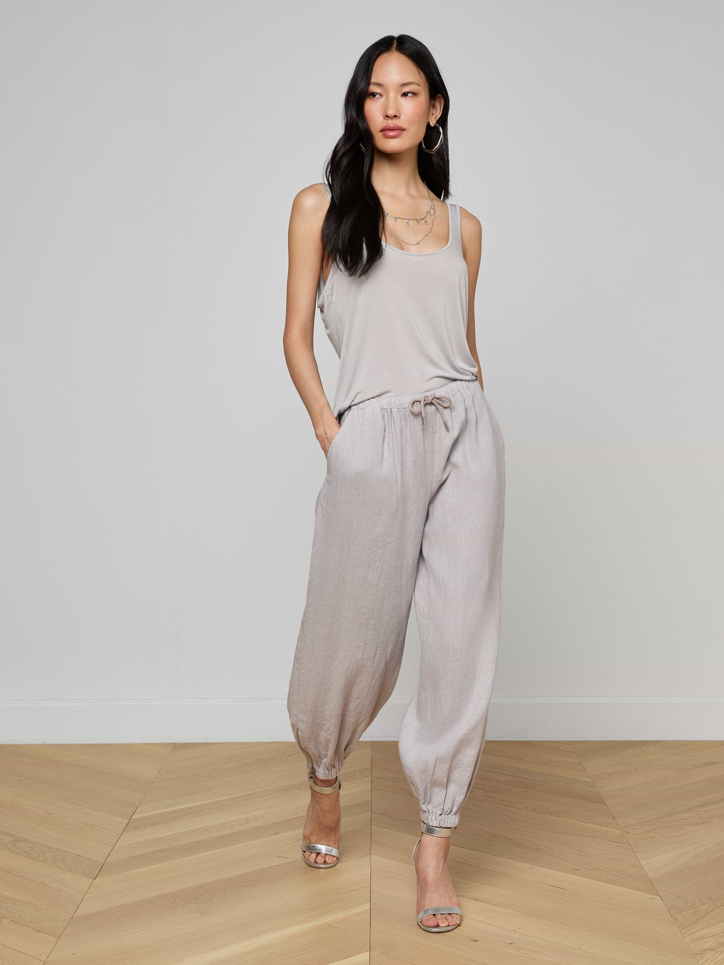 A woman stands against a plain white background, hands in pockets, wearing the Mariko Linen Jogger with a light beige sleeveless top, hoop earrings, and open-toe heels.