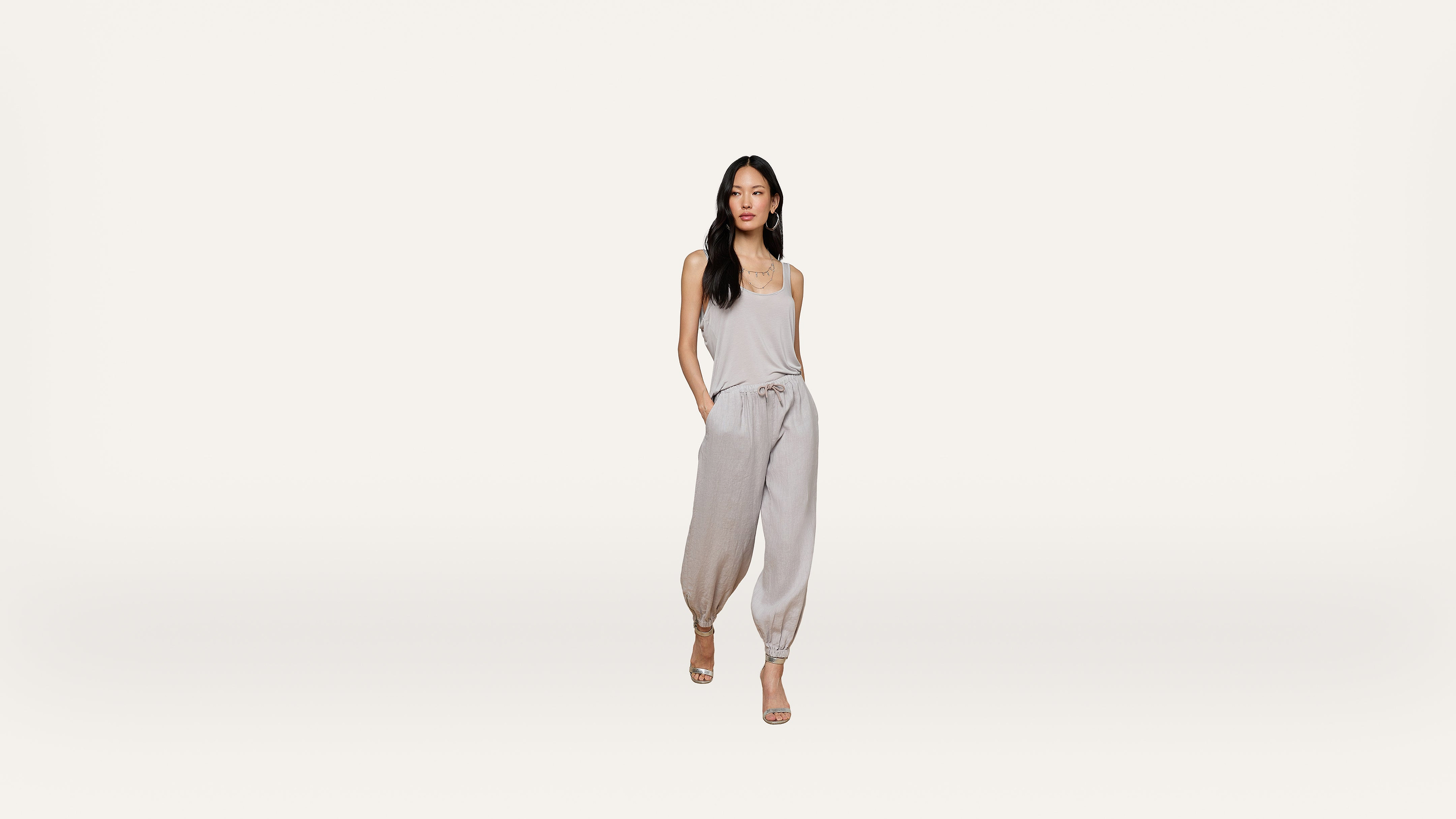 A woman stands against a plain white background, wearing a light gray sleeveless top and matching jogger pants with hands in her pockets, and beige heels.