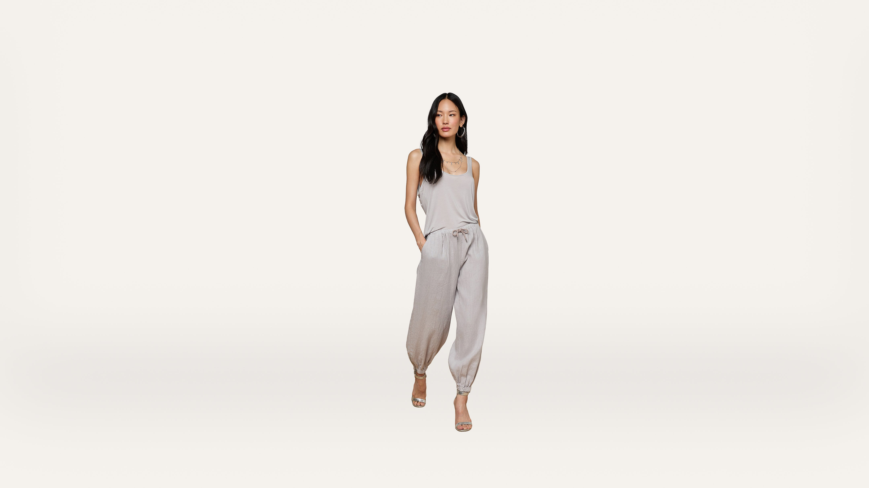 A woman stands against a plain white background, wearing a light gray sleeveless top and matching jogger pants with hands in her pockets, and beige heels.