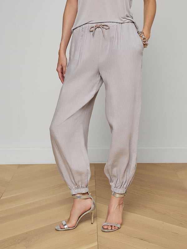 A person is shown from the waist down, modeling the Mariko Linen Jogger in light gray—featuring elastic cuffs and a drawstring waist—paired with a matching top and high-heeled sandals, set against a plain white background.