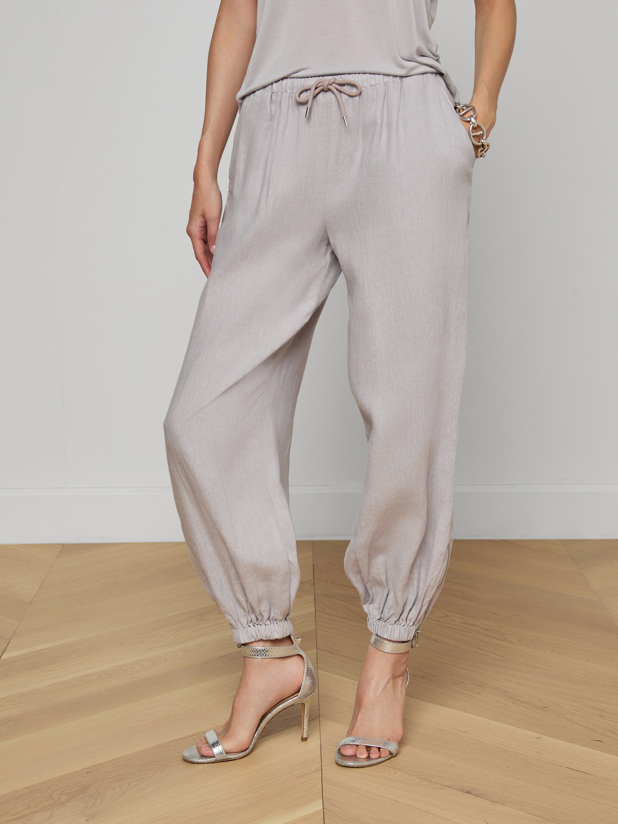 A person is shown from the waist down, modeling the Mariko Linen Jogger in light gray—featuring elastic cuffs and a drawstring waist—paired with a matching top and high-heeled sandals, set against a plain white background.