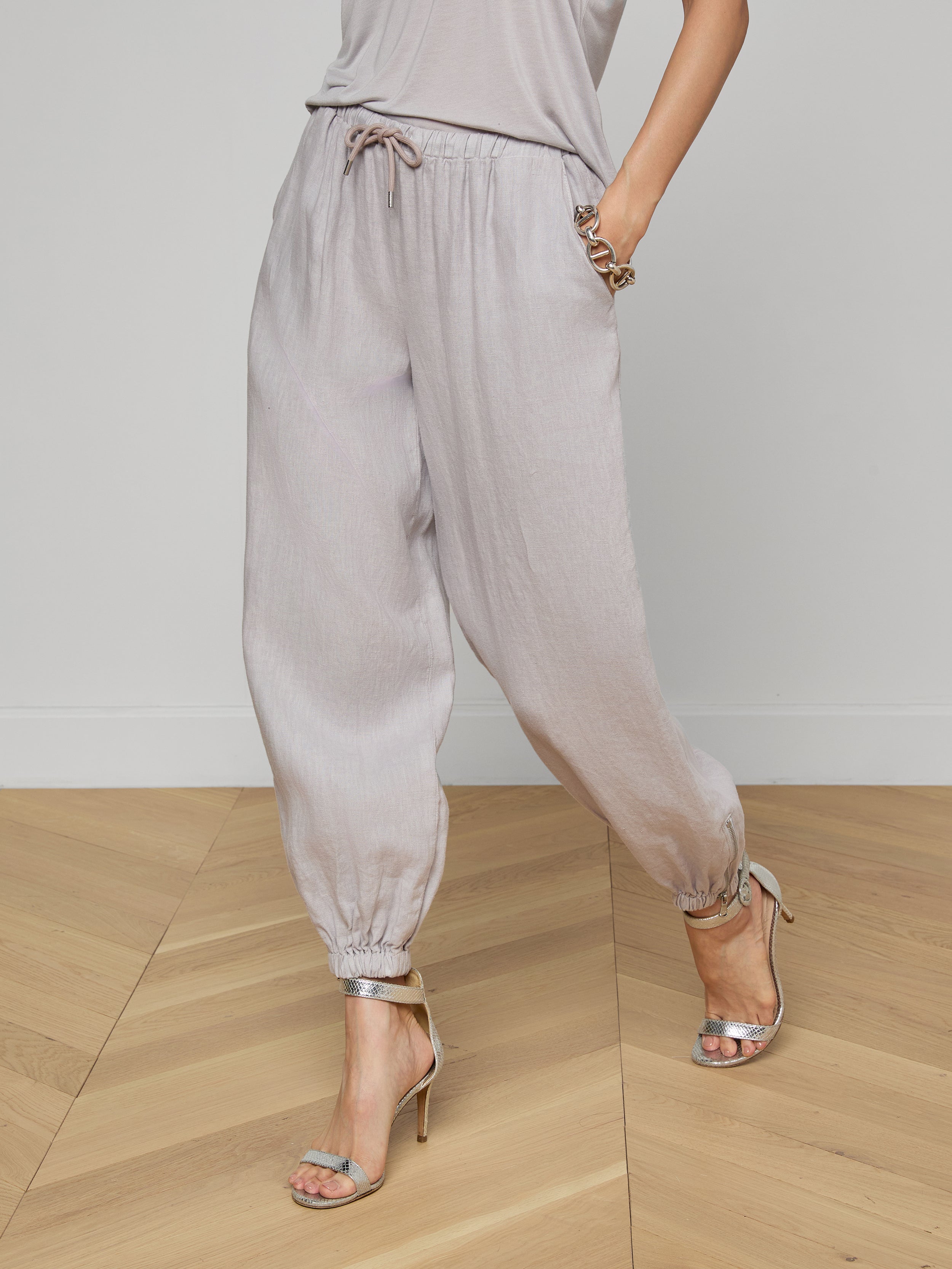Model showcases the Mariko Linen Jogger in light gray, paired with a matching top and styled with silver high-heeled sandals. Posing with hands in pockets against a plain white background, the image highlights only the lower half of the outfit.