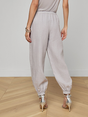 A woman stands on a wooden floor, seen from behind, wearing L’AGENCE Mariko Linen Jogger pants in light gray with elastic ankle cuffs, a taupe top, a silver bracelet, and metallic high-heeled sandals.