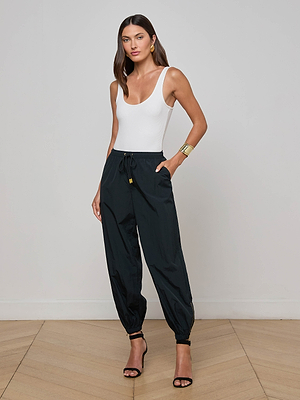 A woman stands indoors against a plain wall, wearing the Mariko Jogger in black with zip cuffs, a white sleeveless bodysuit, black heeled sandals, gold hoop earrings, and a gold cuff bracelet.