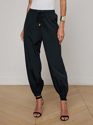 A woman in the Mariko Jogger black drawstring pants, a white sleeveless top, black heeled sandals, and a metallic cuff bracelet stands on a wooden floor against a plain light wall.