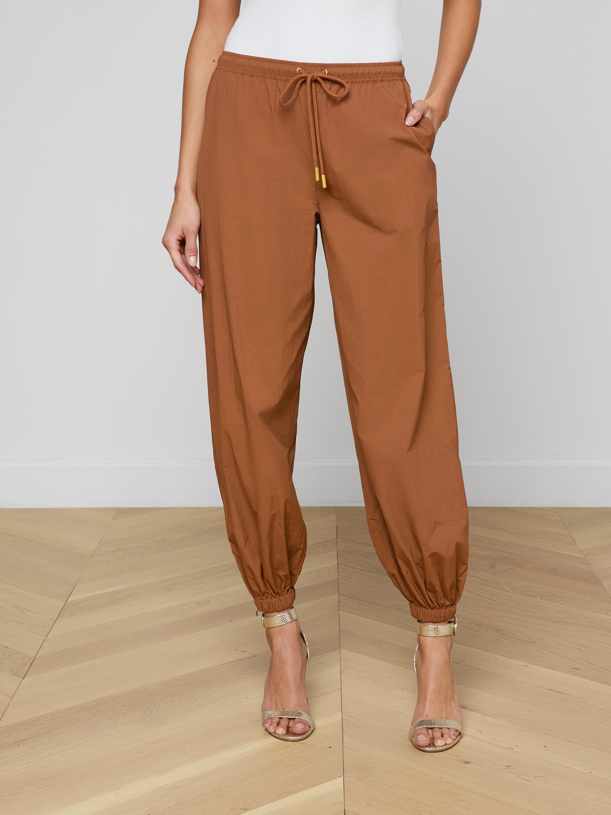 Wearing the Mariko Jogger—modern brown jogger pants crafted from water-repellant nylon with elastic cuffs—paired with beige heeled sandals, a person stands against a plain white background.