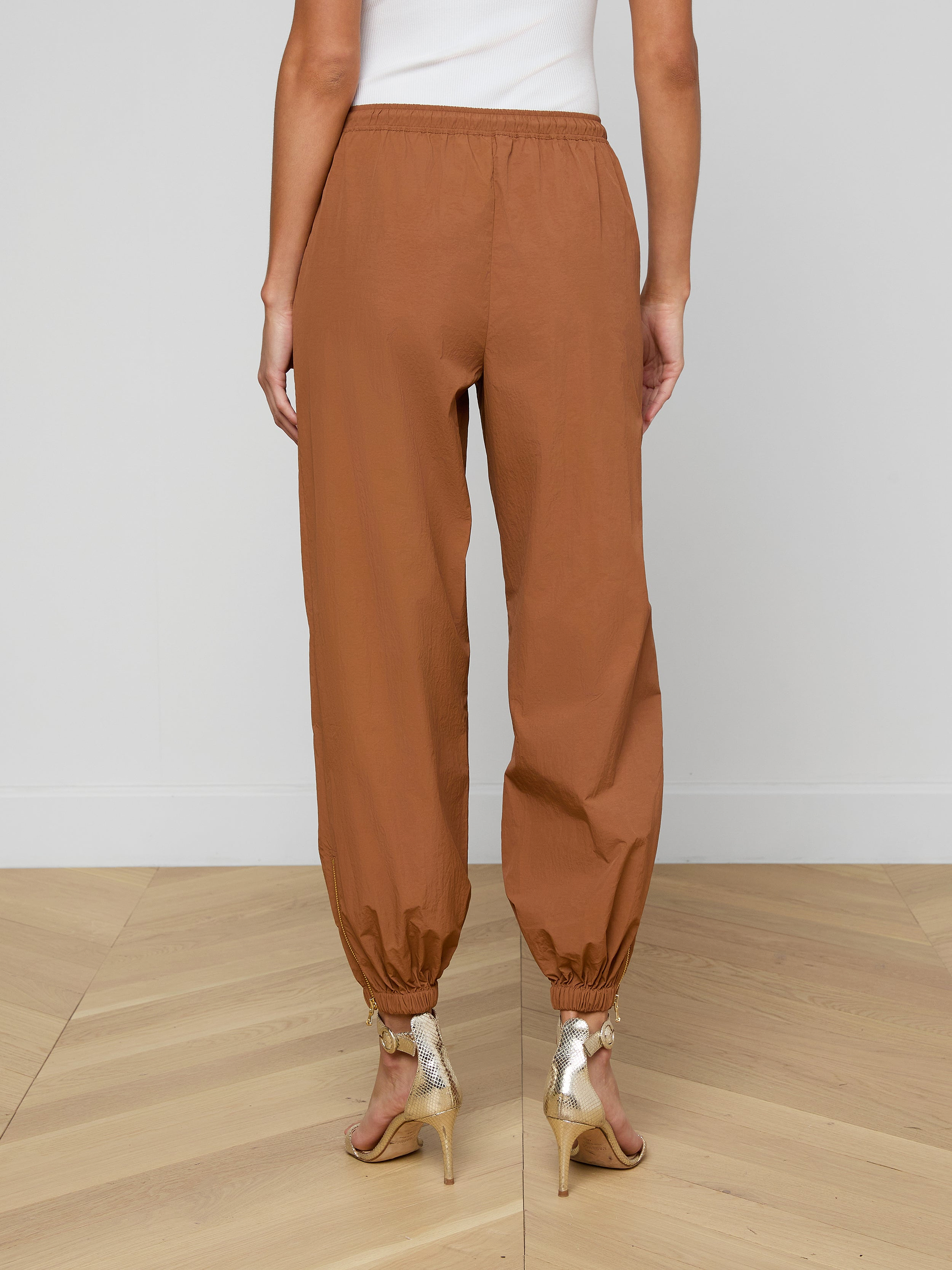 A person is shown from the back against a plain light background wearing the Mariko Jogger—modern brown jogger pants crafted from water-repellent nylon—and beige high-heeled sandals.