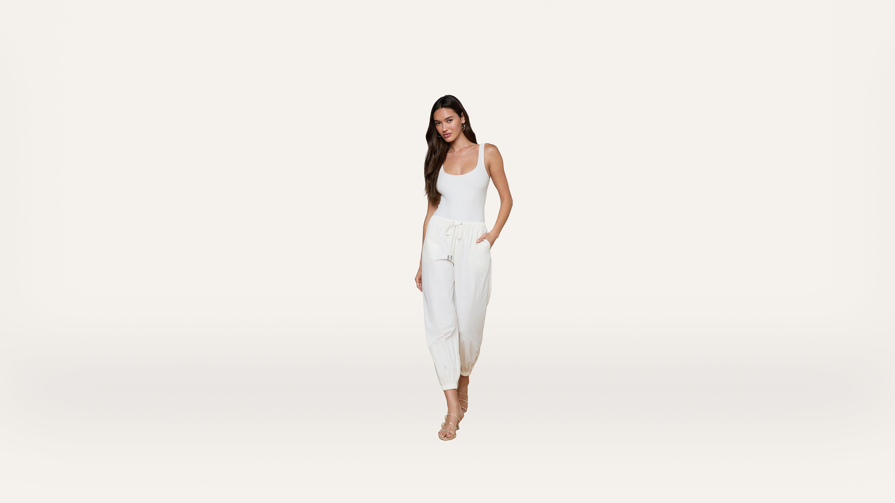 A woman wearing a white tank top and white drawstring pants stands against a plain white background, with her left hand in her pocket and wearing strappy nude heels.