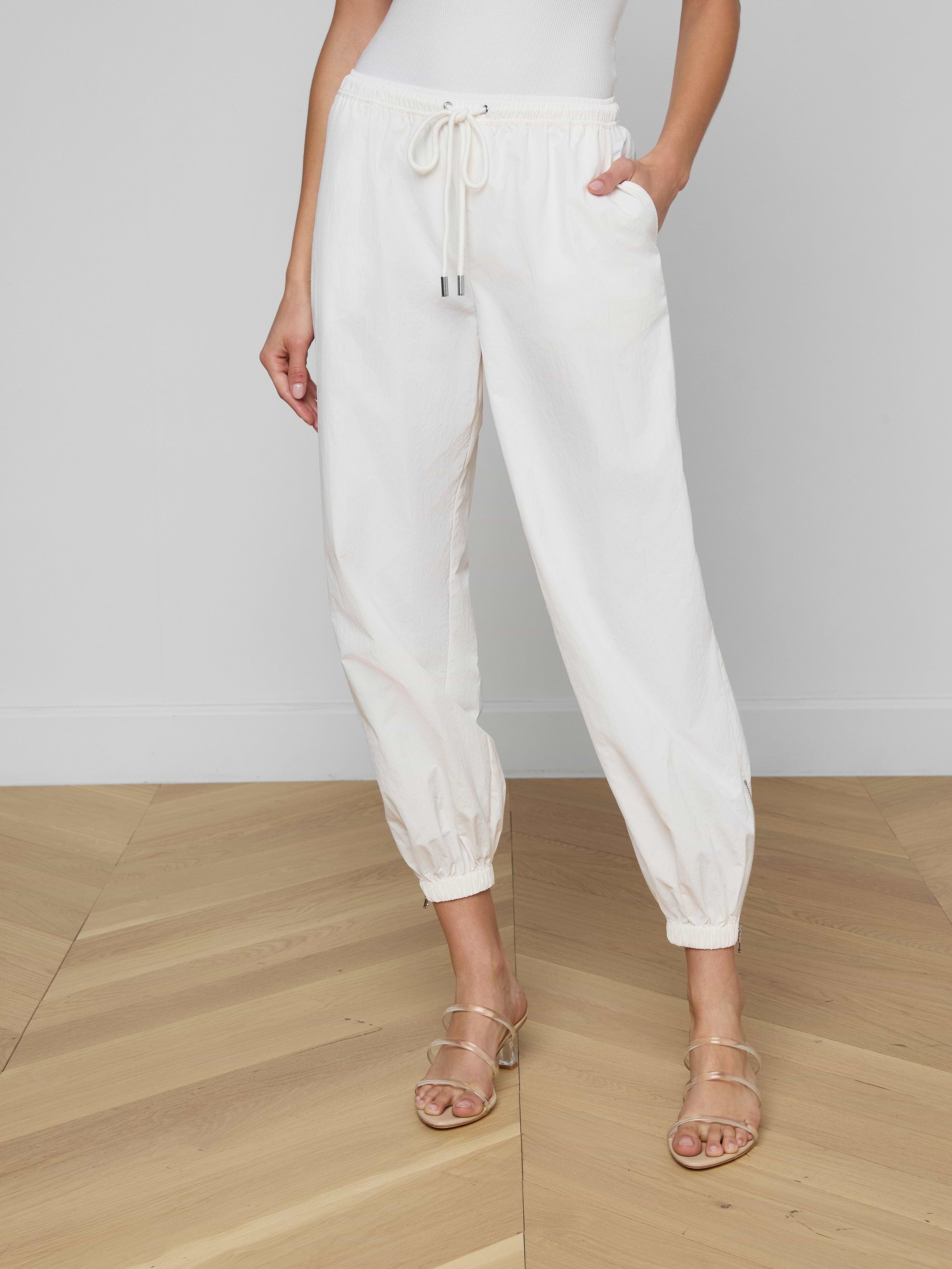 A woman stands against a light background with her hands in her pockets, wearing the Mariko Jogger in modern ivory, crafted from water-repellant nylon. Her beige heeled sandals are visible, while her upper body is mostly out of frame.
