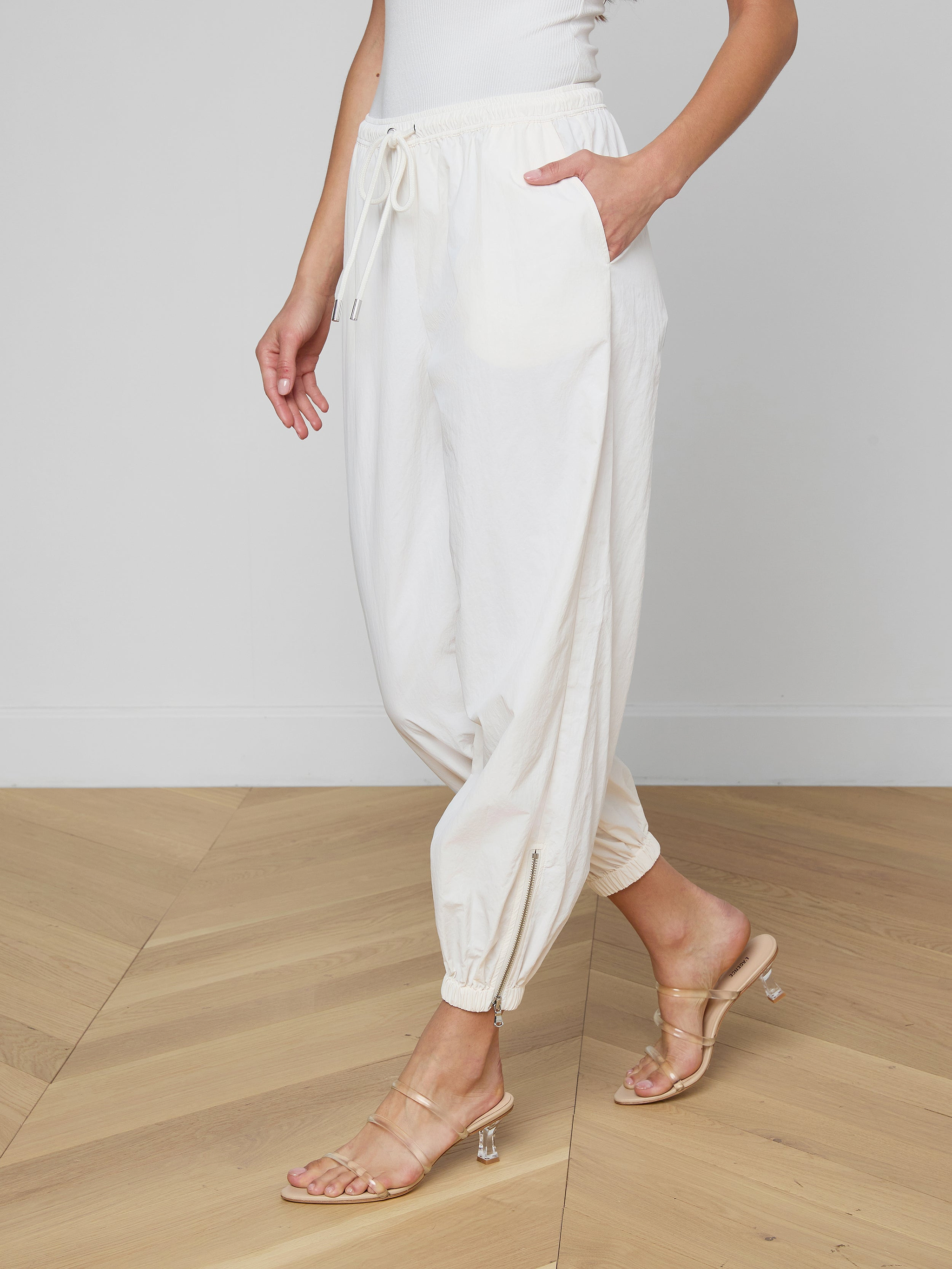 Wearing the Mariko Jogger in ivory—a modern silhouette crafted from water repellent nylon—paired with a white top and beige heeled sandals, a person stands against a plain, light background with one hand in their pocket and their face not visible.