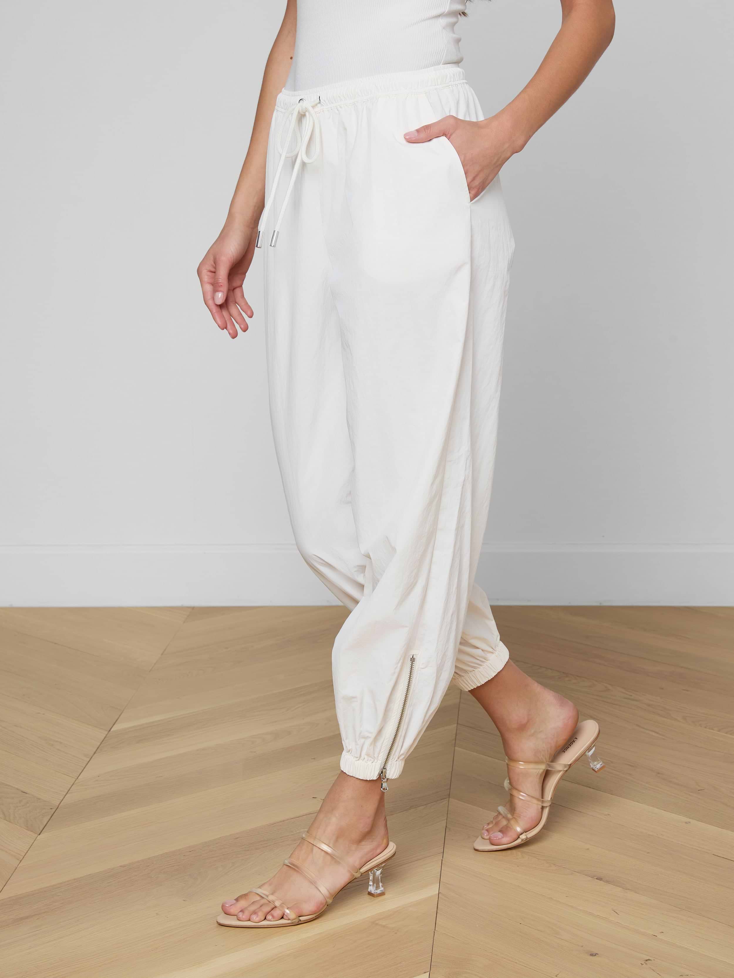 Wearing the Mariko Jogger in ivory—a modern silhouette crafted from water repellent nylon—paired with a white top and beige heeled sandals, a person stands against a plain, light background with one hand in their pocket and their face not visible.