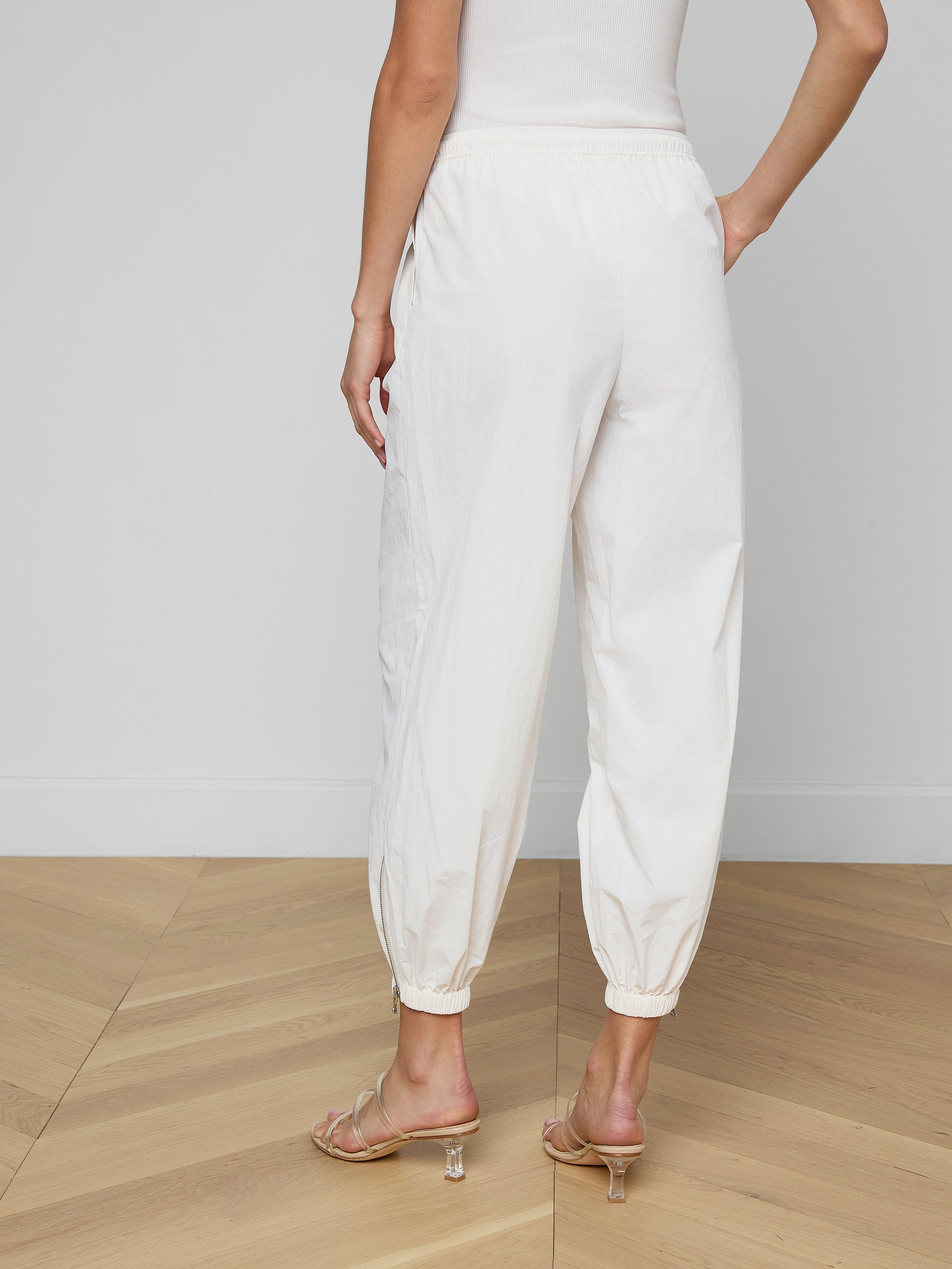 Someone is standing against a plain, light background, shown from the waist down and viewed from behind. They are wearing the Mariko Jogger—modern jogger-style pants in ivory crafted from water-repellent nylon—with heeled sandals.