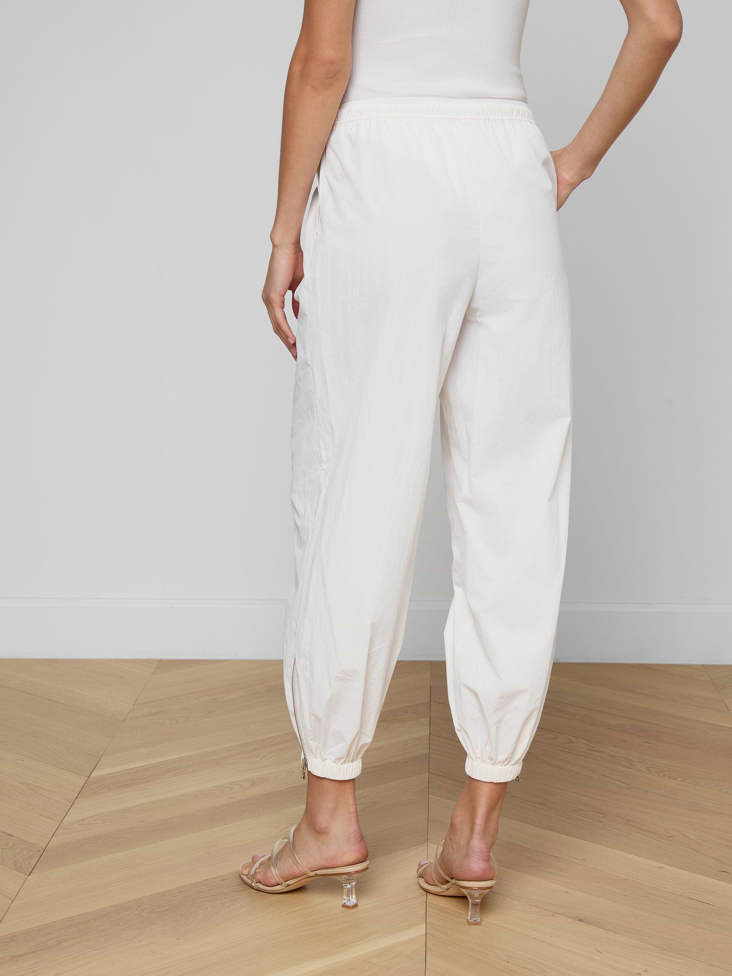 Someone is standing against a plain, light background, shown from the waist down and viewed from behind. They are wearing the Mariko Jogger—modern jogger-style pants in ivory crafted from water-repellent nylon—with heeled sandals.