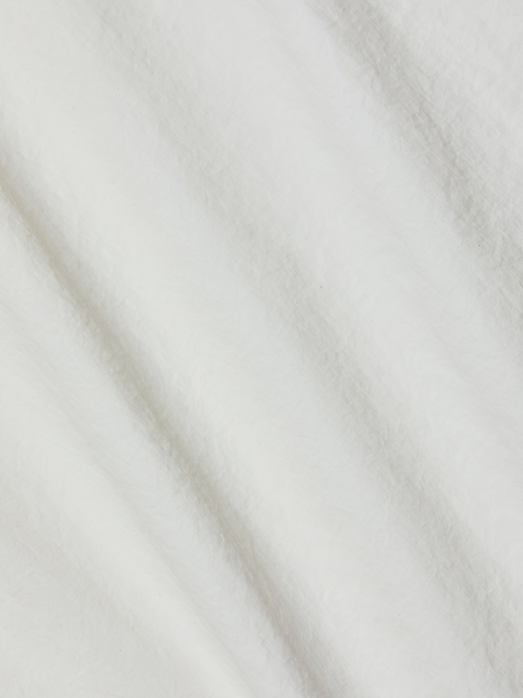 A close-up of the soft, ivory fabric of the Mariko Jogger reveals delicate texture with subtle diagonal folds and shadows. The material looks fluffy and smooth—reminiscent of fleece or cotton, yet crafted from water-repellent nylon for a modern finish.