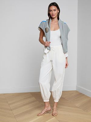 A woman stands in a minimalist room wearing the Mariko Jogger, an ivory tank top, nude sandals, and a light gray jacket draped over her shoulders. She poses with one hand in her pocket.