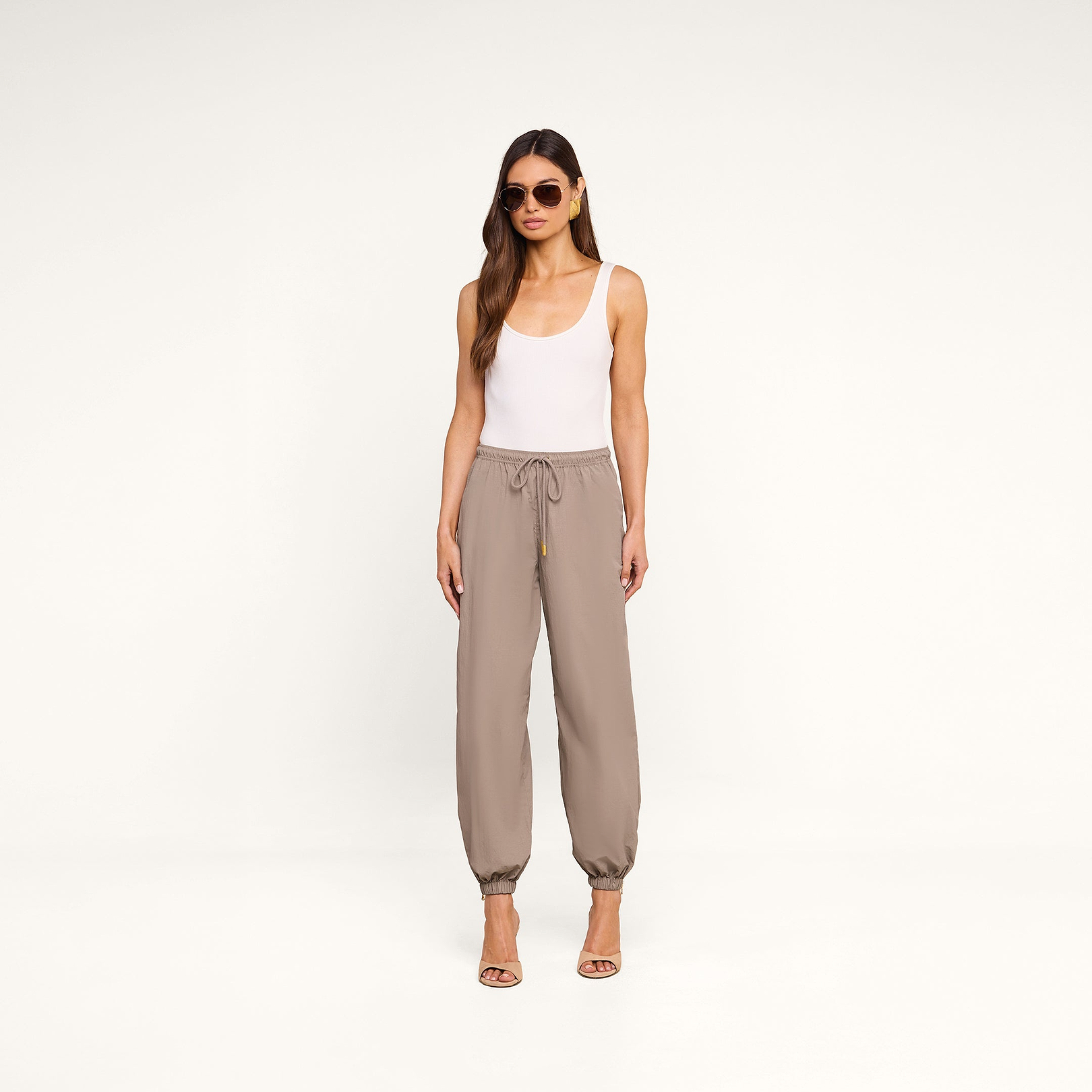A woman stands against a plain white background, wearing sunglasses, a white tank top, high-heeled sandals, and the Mariko Jogger in light cardamom brown—crafted from water-repellent nylon.