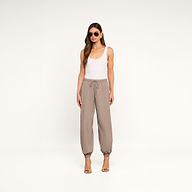 A woman stands against a plain white background, wearing sunglasses, a white tank top, high-heeled sandals, and the Mariko Jogger in light cardamom brown—crafted from water-repellent nylon.