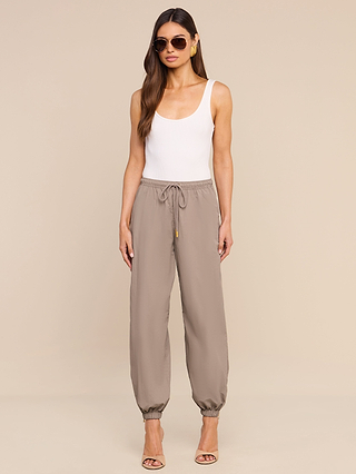 A woman wearing sunglasses, a white tank top, the Mariko Jogger in taupe crafted from lightweight nylon, and beige heels stands against a plain beige background.