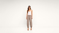 A woman wearing sunglasses, a white tank top, taupe jogger pants, and heeled sandals stands against a plain white background.