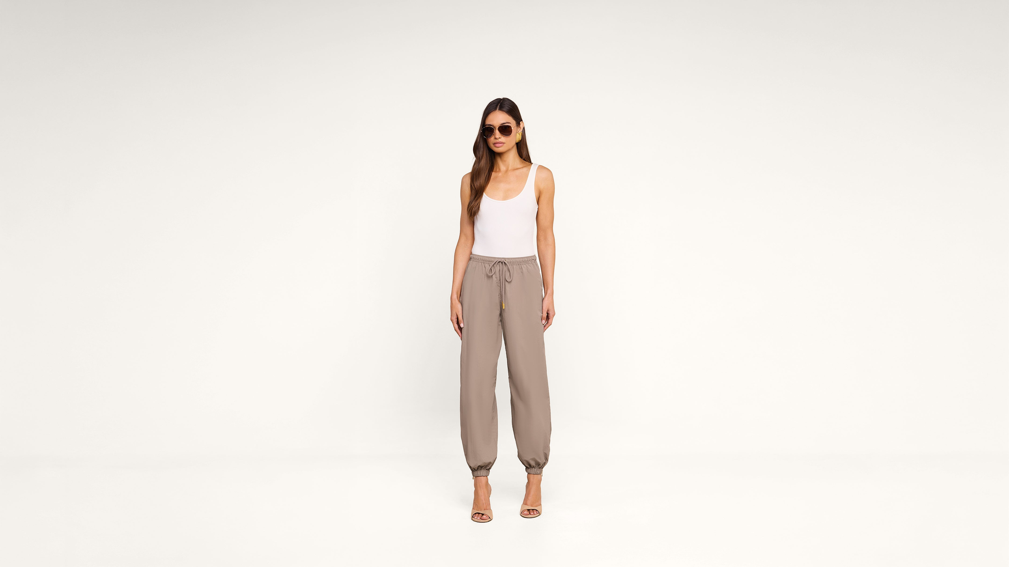 A woman wearing sunglasses, a white tank top, taupe jogger pants, and heeled sandals stands against a plain white background.