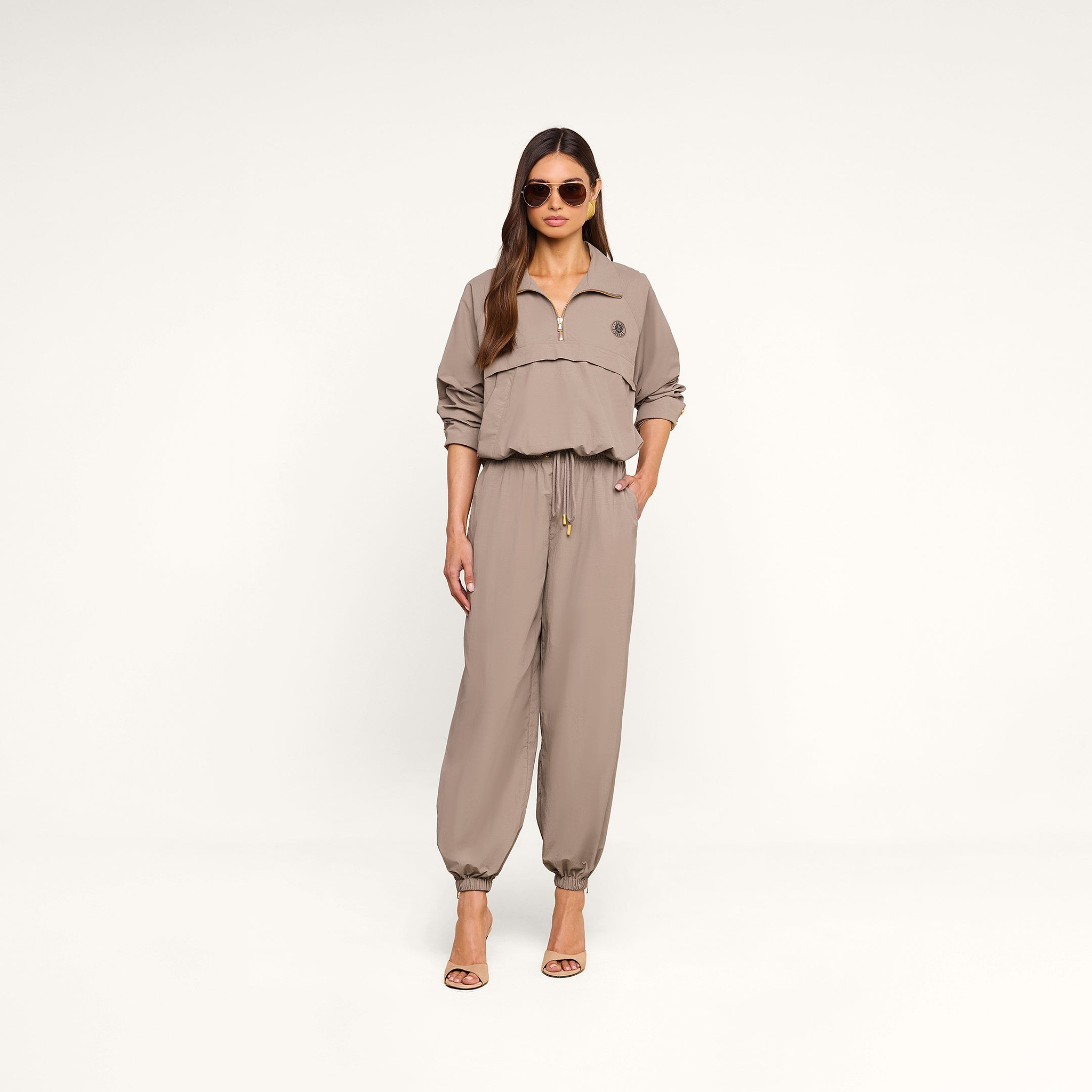 A woman wearing sunglasses and a light cardamom brown, long-sleeve zip-up top with matching Mariko Jogger pants crafted from water repellent nylon and heeled sandals stands against a plain white background.