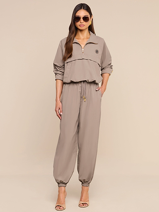 Woman in a tan Mariko Jogger zip-up long sleeve jumpsuit crafted from lightweight, water repellent nylon with a drawstring waist and cuffed ankles, wearing sunglasses and heels, standing against a beige background. Inspired by the signature Mariko Jogger silhouette, this piece perfectly balances style and function.