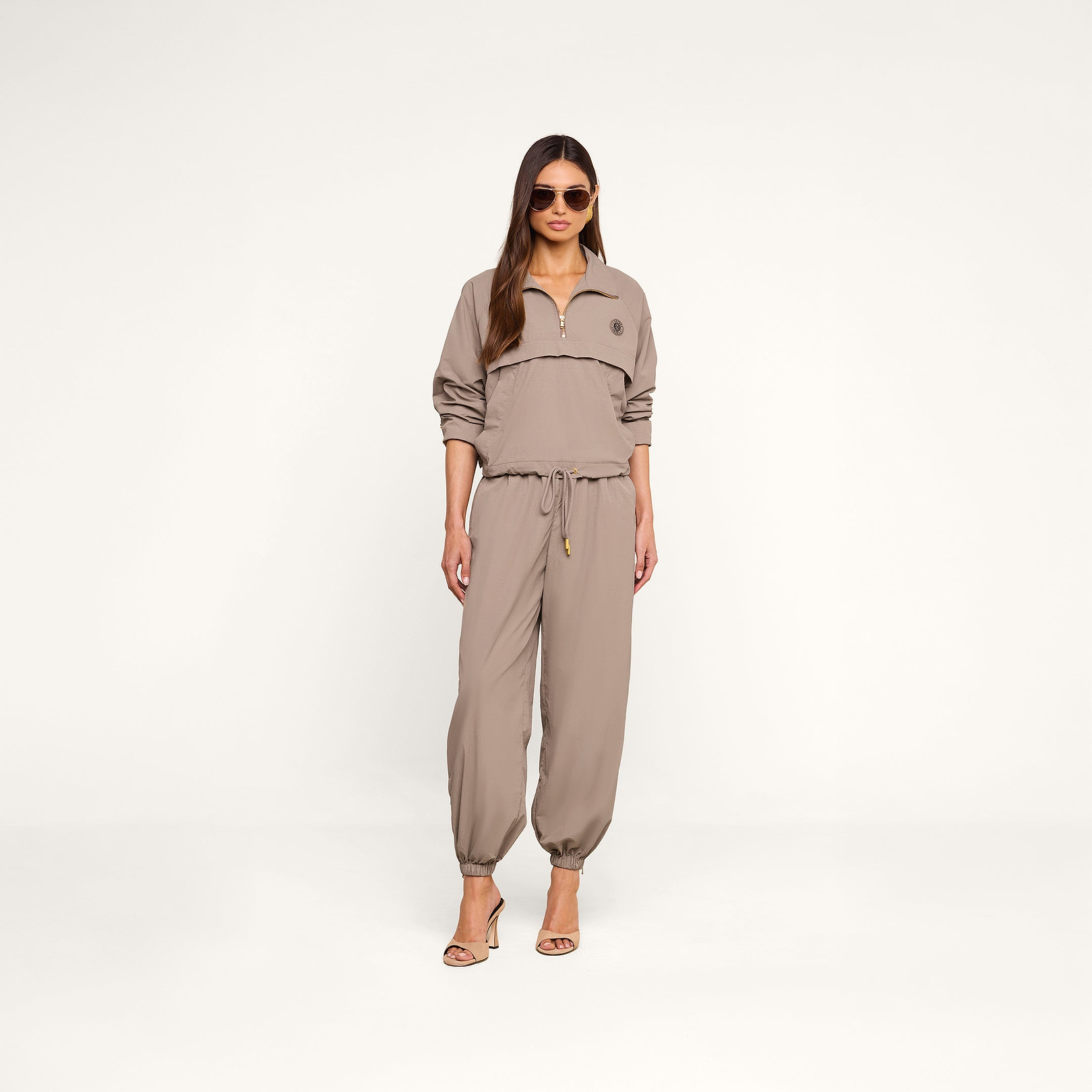 A woman stands in heels against a plain white background, wearing sunglasses and the Mariko Jogger—a light cardamom brown jumpsuit crafted from water-repellent nylon, featuring a zip-up top and drawstring waist.
