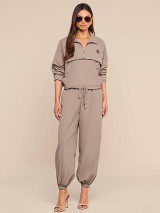 A woman stands against a beige background in heeled sandals, wearing taupe sunglasses, a taupe half-zip pullover, and the Mariko Jogger—lightweight, water-repellent nylon pants featuring a drawstring waistband.