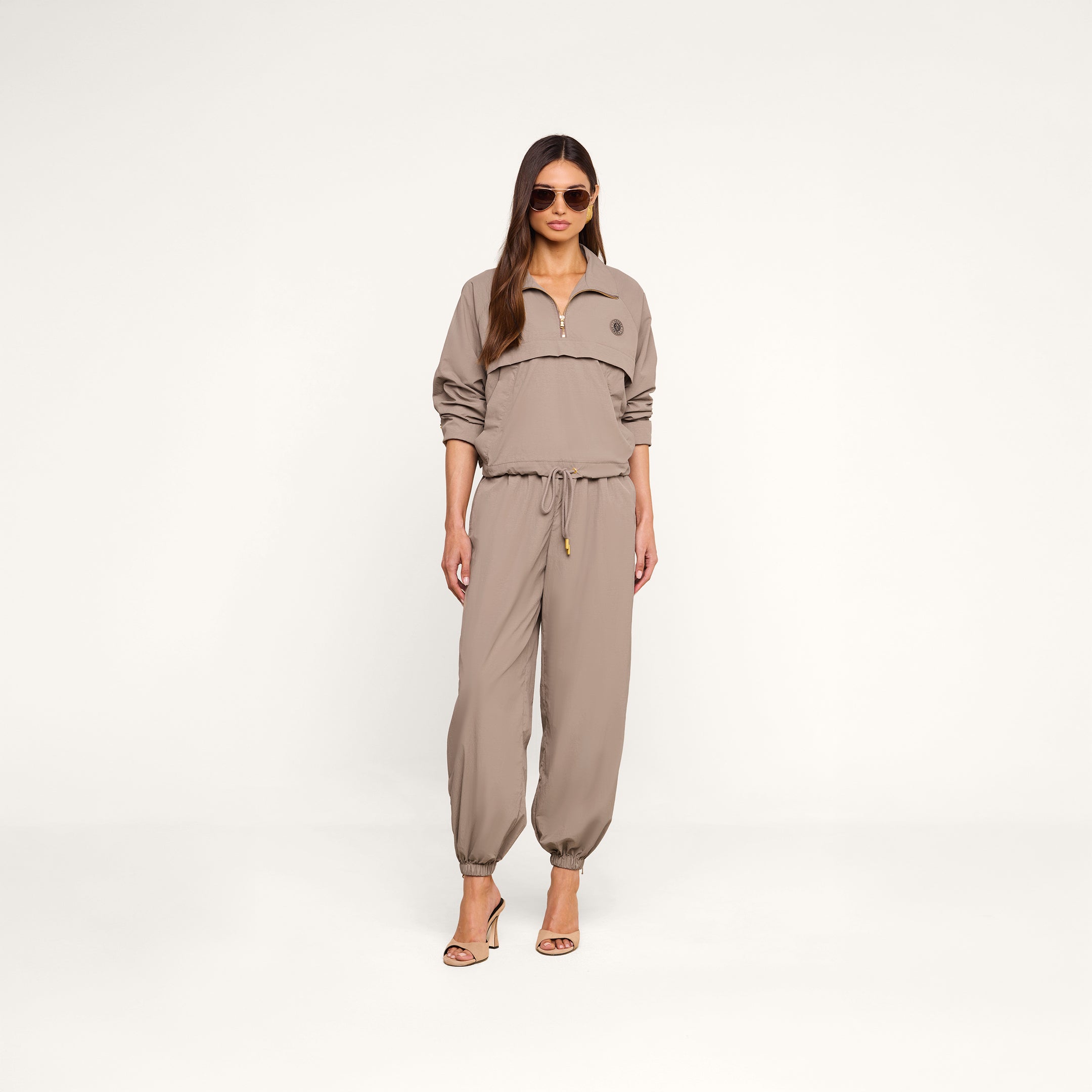 A woman stands in heels against a plain white background, wearing sunglasses and the Mariko Jogger—a light cardamom brown jumpsuit crafted from water-repellent nylon, featuring a zip-up top and drawstring waist.