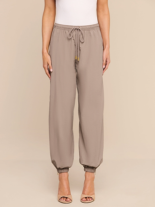 Clad in the Mariko Jogger—taupe pants crafted from lightweight nylon with elastic cuffs—a person pairs them with beige heels and stands against a plain beige background.