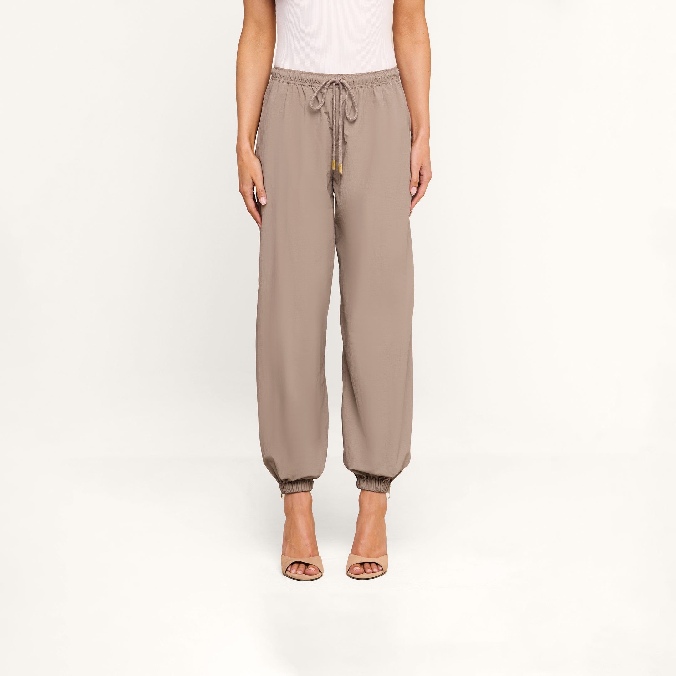 Wearing the Mariko Jogger in light cardamom brown featuring an elastic waistband and drawstring, paired with a white top and nude open-toe heels, a person stands against a plain white background.