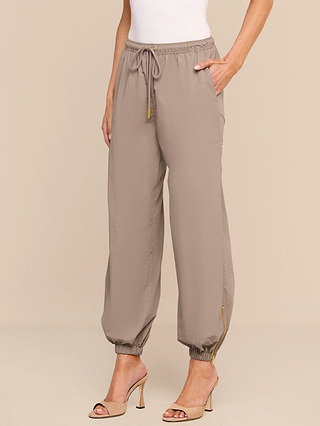 Wearing the Mariko Jogger, crafted from lightweight nylon with elastic cuffs and gold ankle zippers, styled with a white sleeveless top and beige high-heeled sandals.