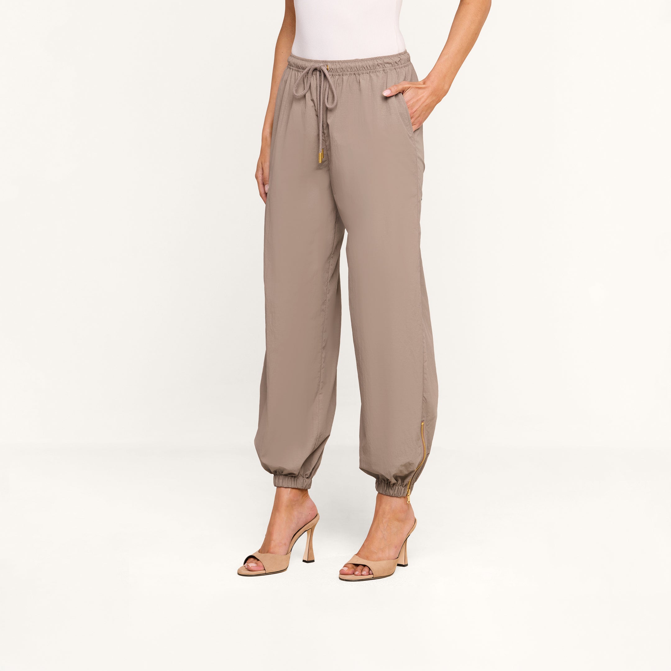 A woman stands against a plain white background wearing the Mariko Jogger in light cardamom brown, featuring a drawstring waist and pockets, paired with a white top and beige high-heeled sandals.