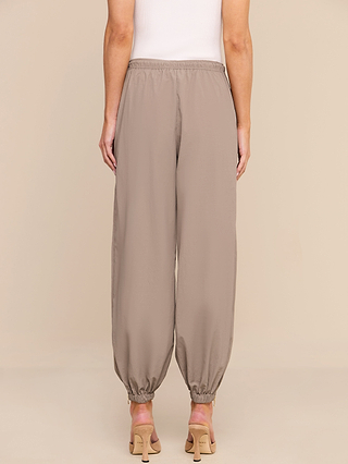 A person is seen from the back, standing against a neutral background, wearing the Mariko Jogger in taupe—crafted from lightweight nylon with an elastic waistband and ankle cuffs—paired with a white sleeveless top and beige heeled sandals.