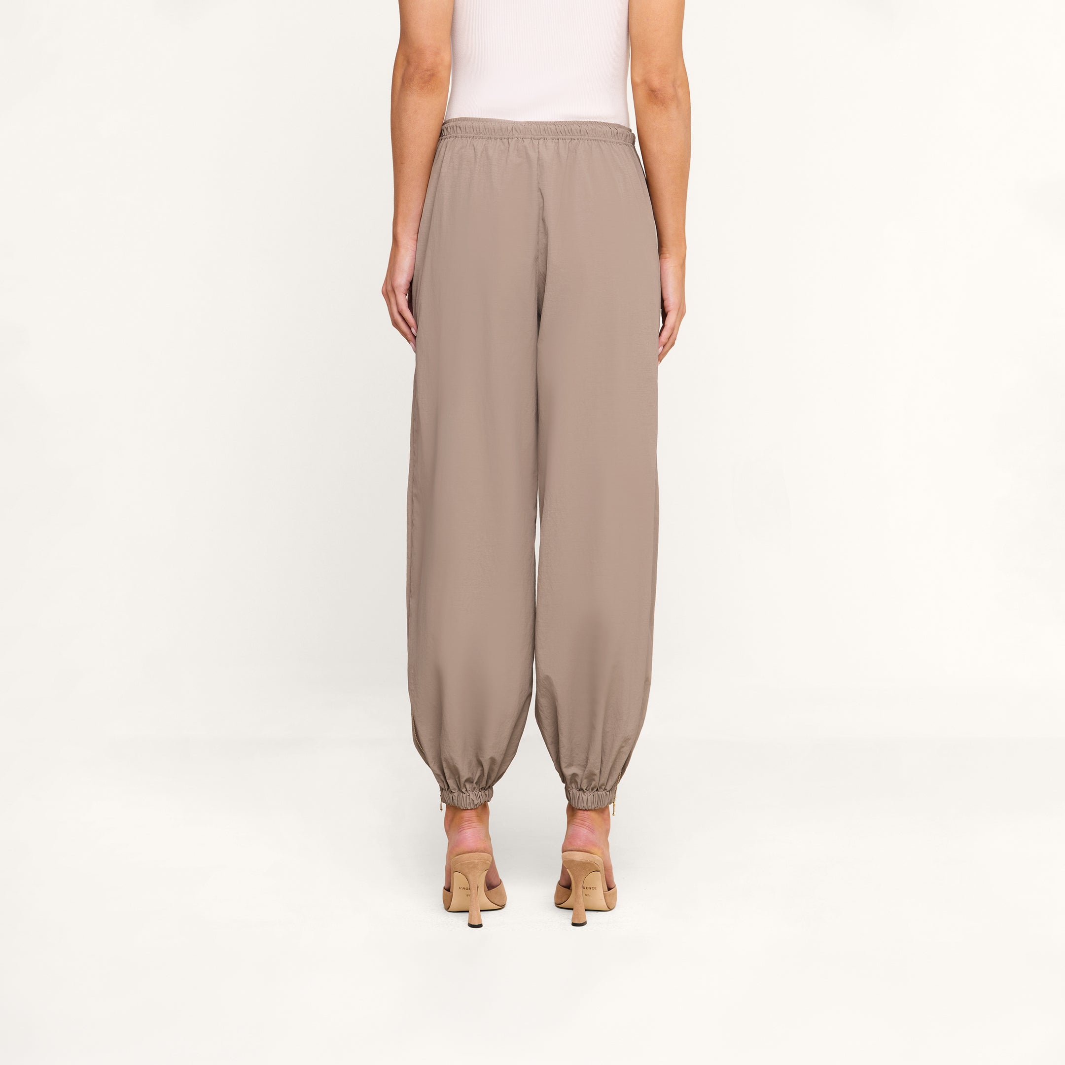 A person wearing Mariko Jogger pants in light cardamom brown and heeled sandals stands facing away, hands at their sides, against a plain white background.