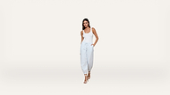 Woman in a white tank top, light blue jogger pants, and white heeled sandals stands with one hand in her pocket against a plain, light background.