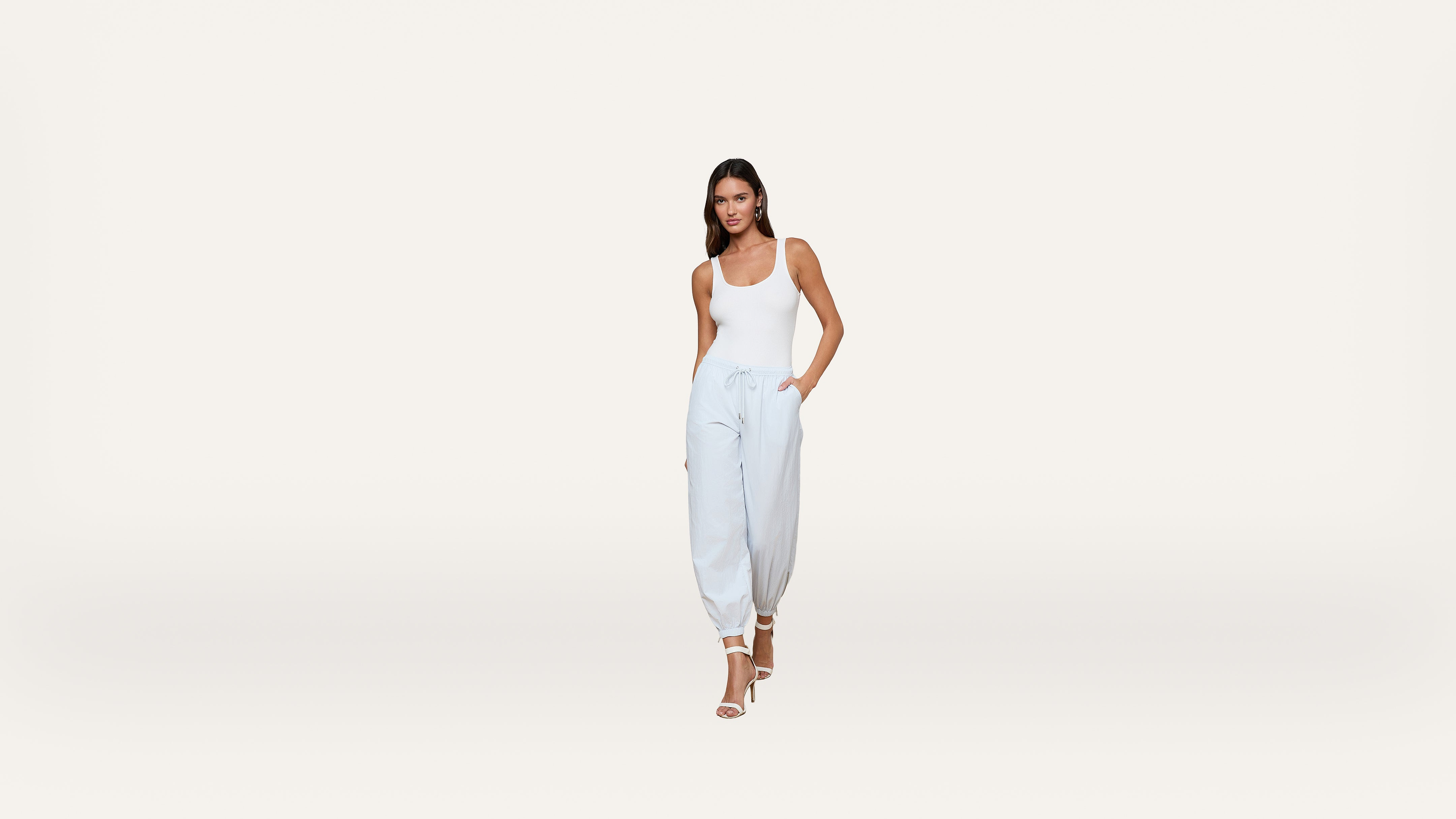 Woman in a white tank top, light blue jogger pants, and white heeled sandals stands with one hand in her pocket against a plain, light background.
