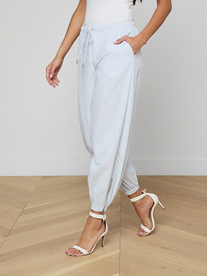 A woman wears the Mariko Jogger in light blue with a drawstring waist and side pockets, paired with white heeled sandals, standing on a wooden floor against a plain white wall.