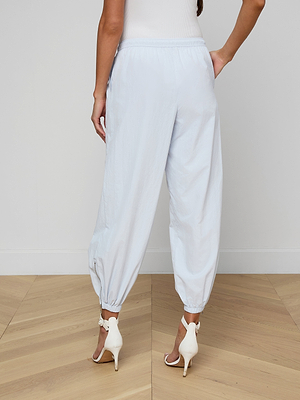 A woman stands with her back to the camera, wearing the Mariko Jogger in light blue, a white sleeveless top, and white high-heeled sandals on a light wooden floor.