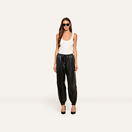 A woman wearing sunglasses, a white tank top, the Mariko Leather Jogger pants, and black heels stands facing forward against a plain light background.