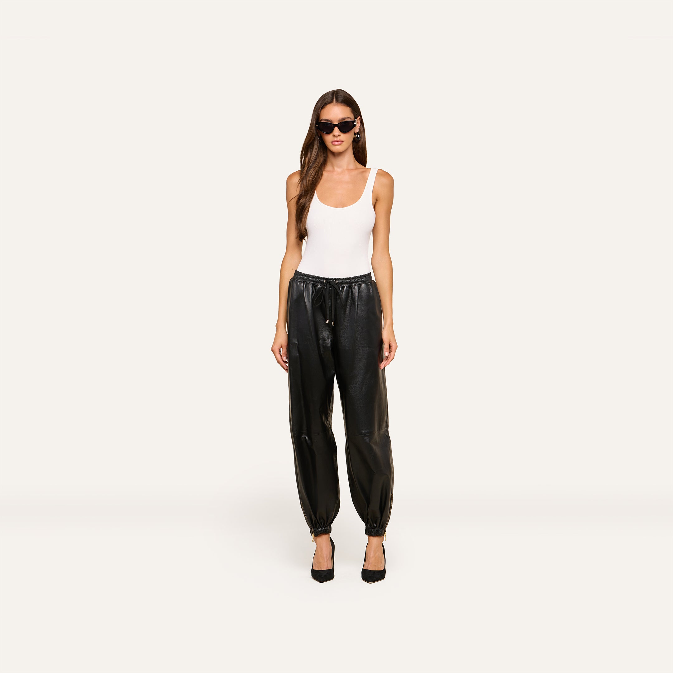 A woman wearing sunglasses, a white tank top, the Mariko Leather Jogger pants, and black heels stands facing forward against a plain light background.