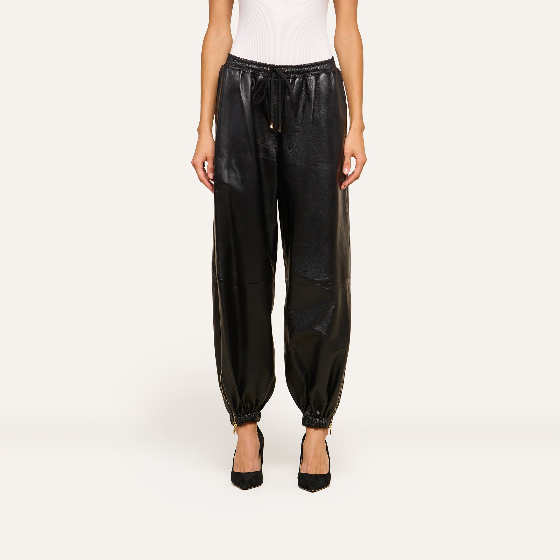 Wearing the Mariko Leather Jogger in black lambskin—with a drawstring waist and elasticated ankles—a person pairs the joggers with a white sleeveless top and black pointed-toe heels, standing against a light background.