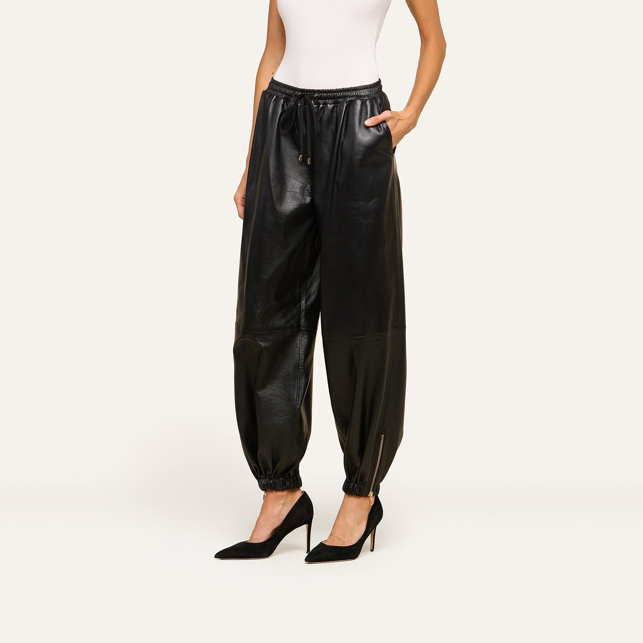 Wearing the Mariko Leather Jogger crafted from black faux leather with elastic cuffs and a drawstring waist, the woman pairs it with a white sleeveless top and black pointed-toe heels, standing stylishly with one hand in her pocket.