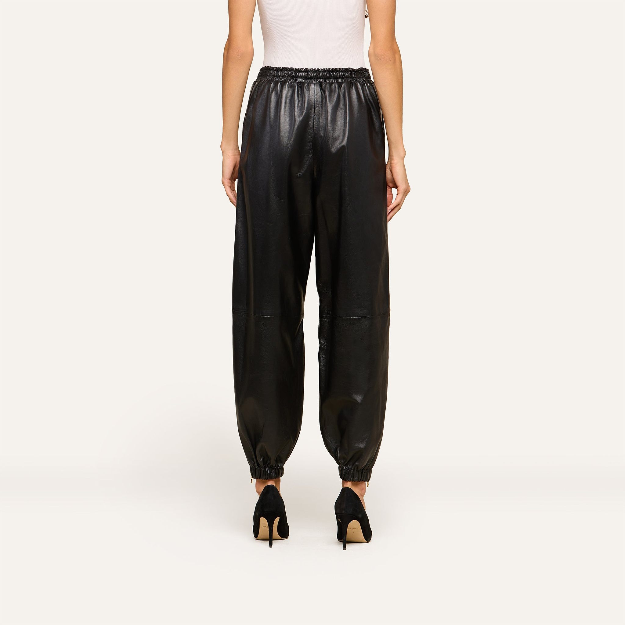 A person wearing a white sleeveless top and the Mariko Leather Jogger in black lambskin with elastic ankles, standing and facing away against a plain, light-colored background.