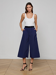A woman stands against a plain light background wearing a white tank top, the Shoda Cropped Wide-Leg Pant in navy blue with a black belt, and black high-heeled sandals.