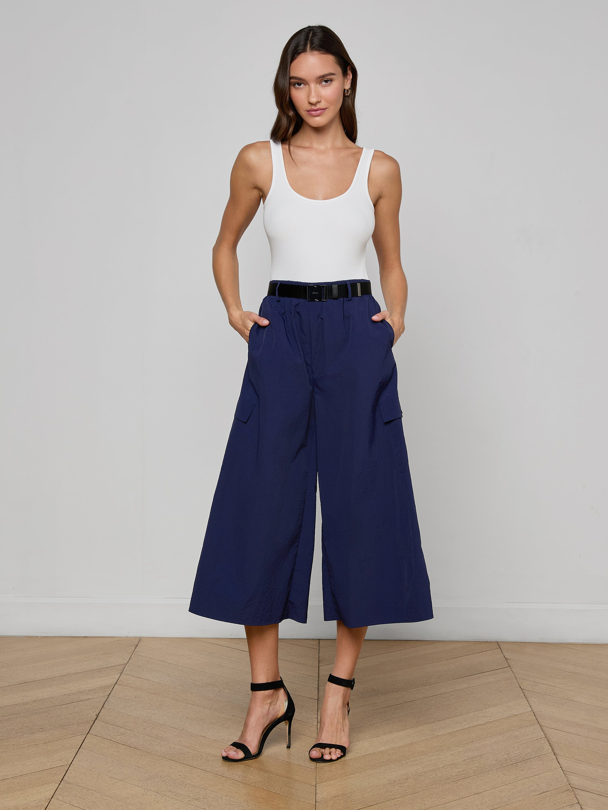 A woman stands against a plain light background wearing a white tank top, the Shoda Cropped Wide-Leg Pant in navy blue with a black belt, and black high-heeled sandals.