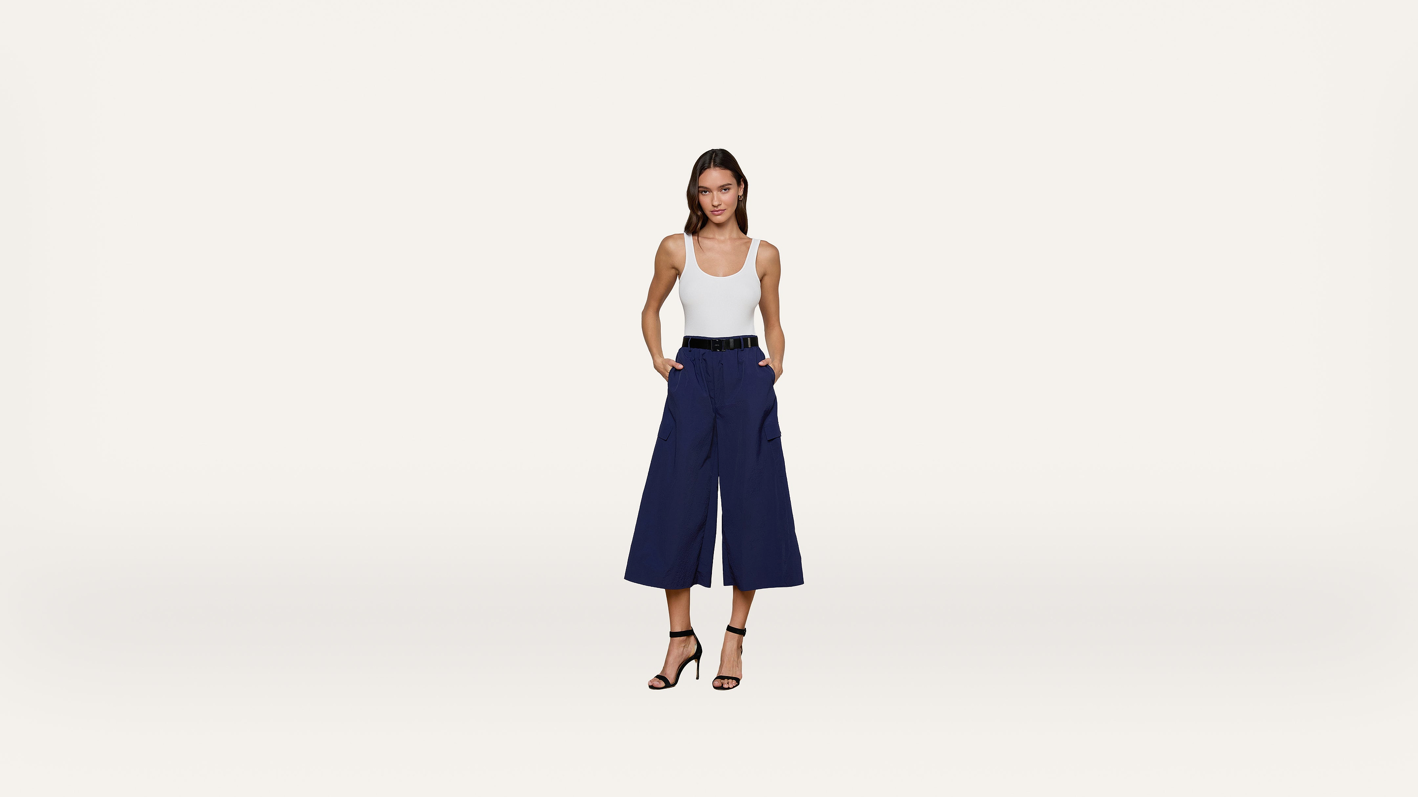 A woman stands against a white background, wearing a white tank top, navy wide-leg cropped pants, black belt, and black high-heeled sandals, with hands in her pockets.