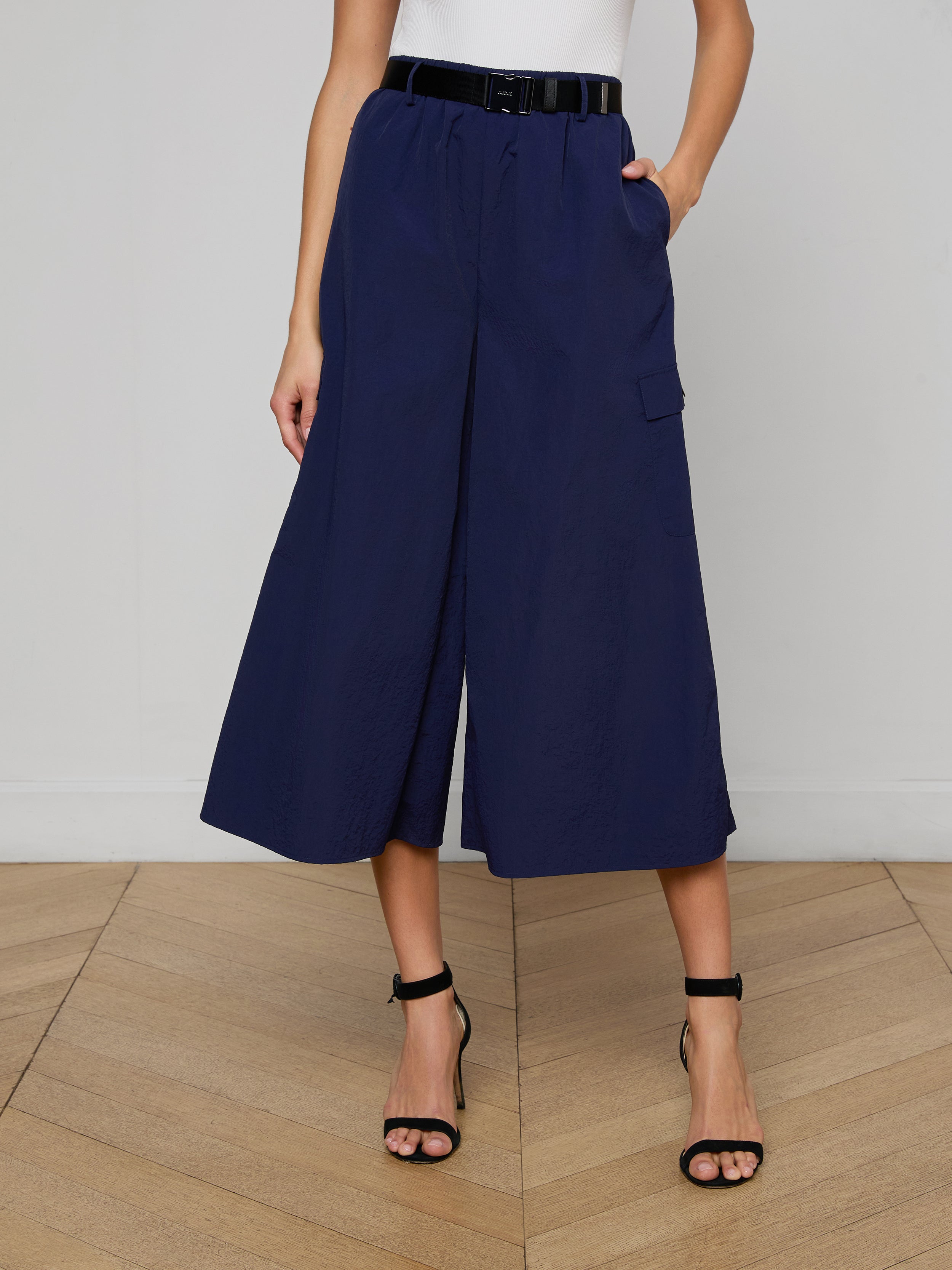 A woman models the Shoda Cropped Wide-Leg Pant in navy blue, styled with a black belt and black high-heeled sandals. Her hands are tucked into the pants’ pockets, while her upper body remains out of frame against a plain white background.